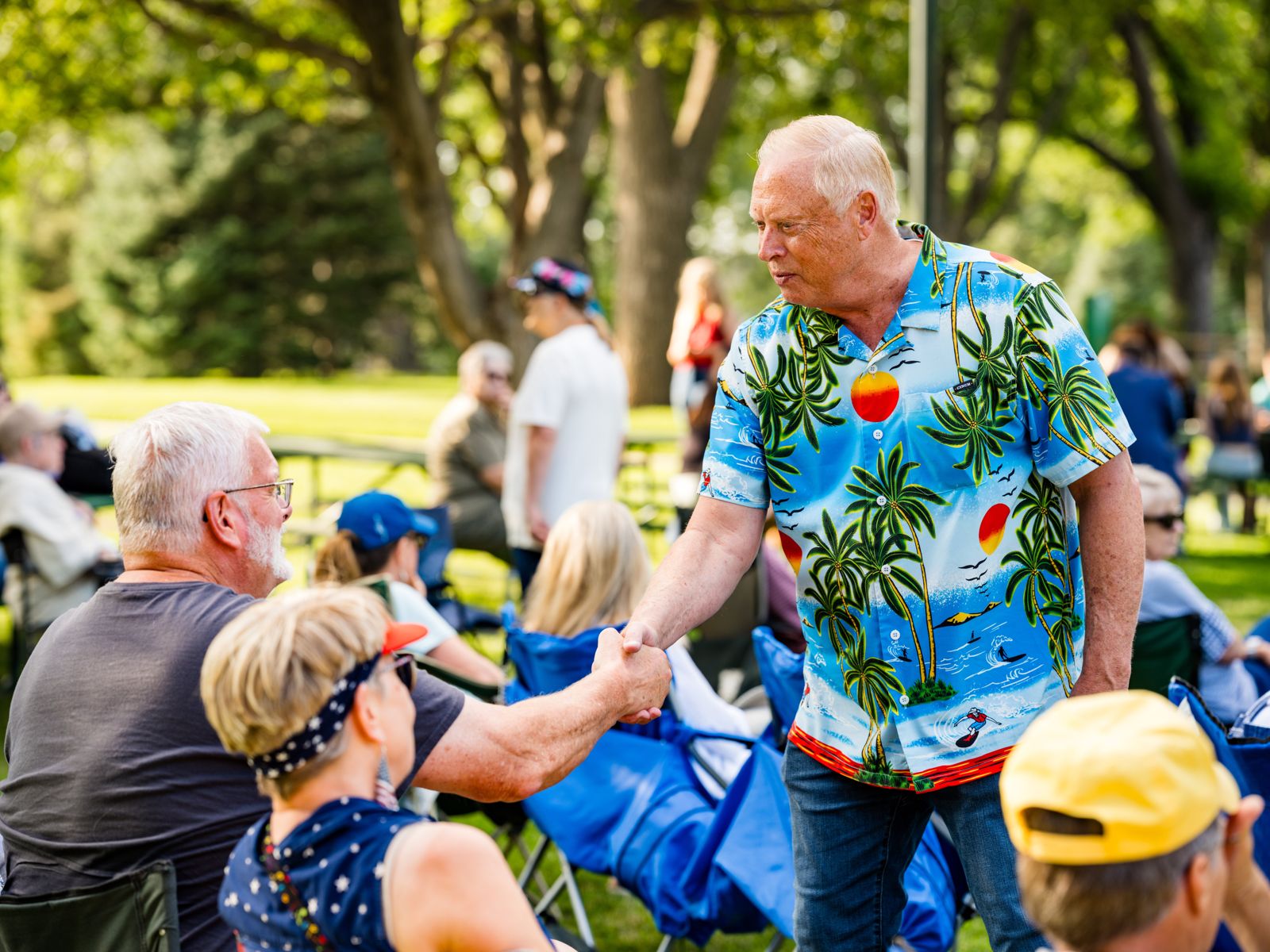 A neighbor in a palm-tree shirt shaking hands with picnic guests in lawn chairs